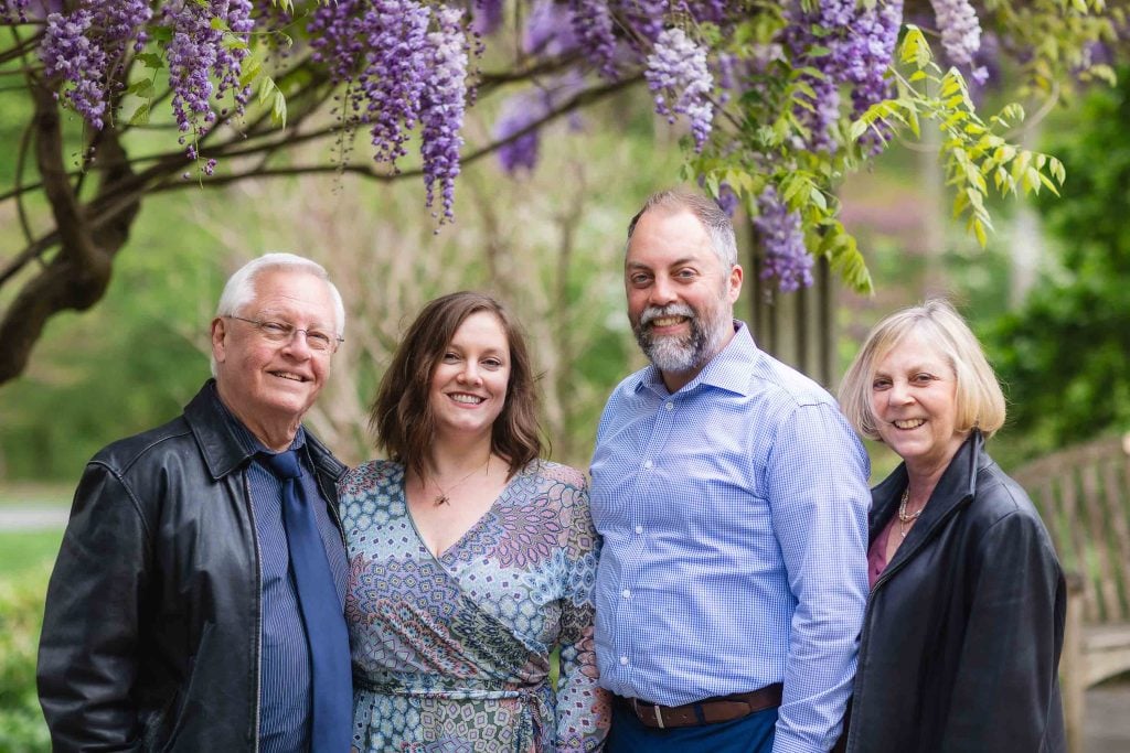 Four people stand smiling under a wisteria tree in what appears to be an extended family portrait. The group includes two men and two women, all dressed in casual to semiformal attire. A wooden bench is partially visible in the background.