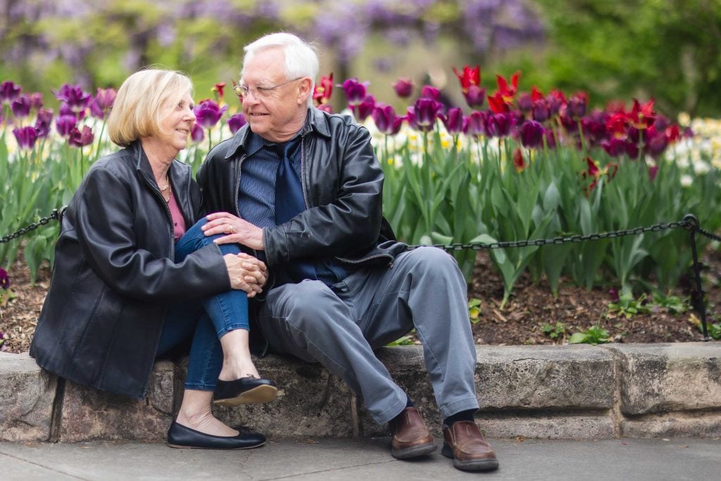 An older couple sitting on a low stone wall in front of a flower bed with red and purple tulips at Brookside Gardens, smiling and holding hands, creating a charming portrait.