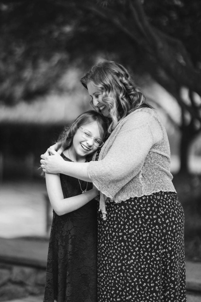 A woman and a young girl share a hug and smile warmly in a black and white family portrait set in the picturesque Brookside Gardens.