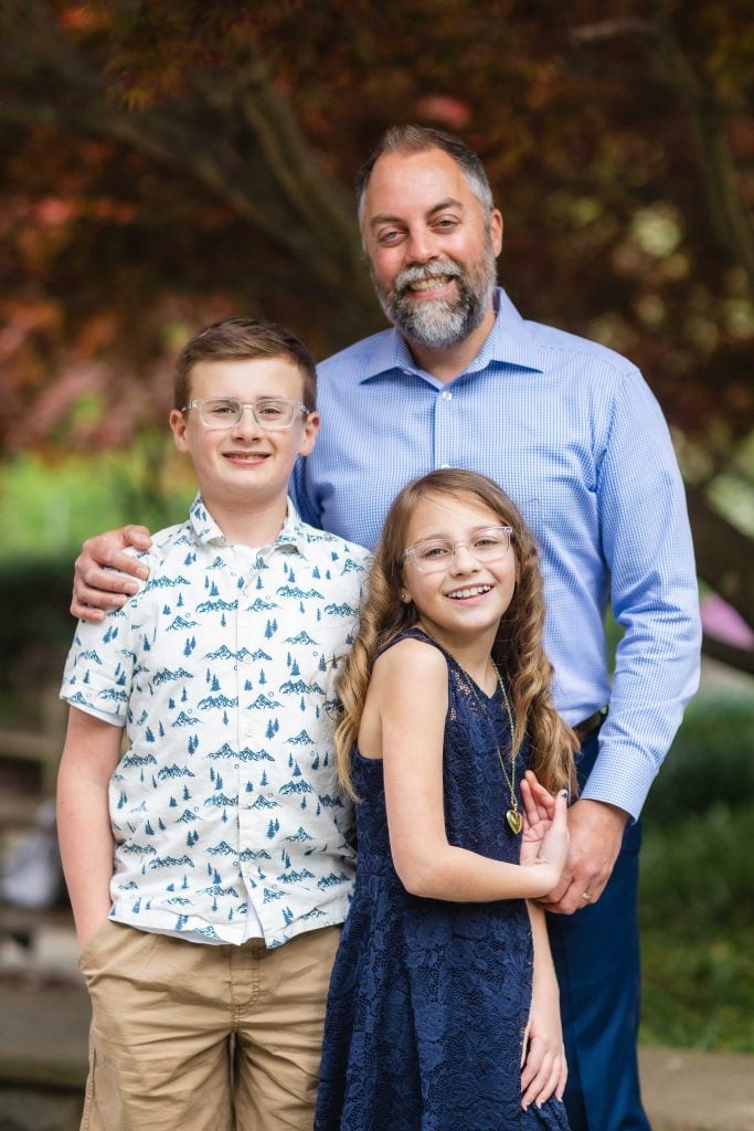 A man with a beard stands behind two smiling children, one boy and one girl, outdoors at Brookside Gardens. The boy wears a patterned shirt and khaki shorts, while the girl wears a blue lace dress. This lovely family moment captures their joy amidst the lush greenery.