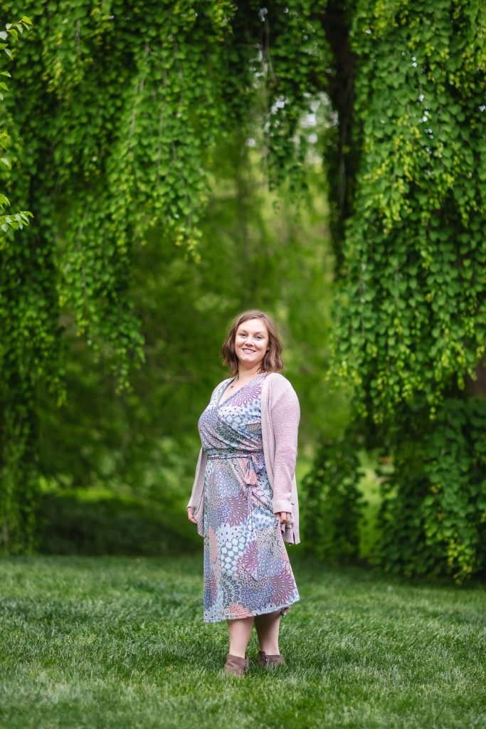 A woman in a patterned dress and pink cardigan stands on the grass at Brookside Gardens, smiling at the camera, with lush, green hanging foliage providing a stunning backdrop for this extended portrait.