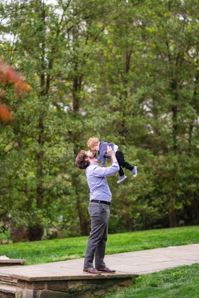 A joyful portrait captures a man lifting a child into the air while standing on a stone pathway in an enchanting garden with lush green trees in the background, celebrating moments of extended family love.