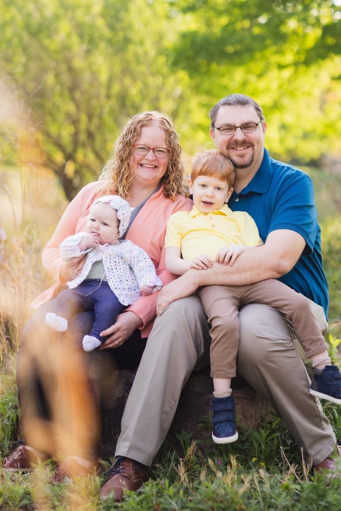 A man and woman sit on a rock, each holding a child in Quiet Waters Park. The woman cradles a baby in a white sweater, while the man holds a boy in a yellow shirt. Portraits of serene family moments are framed by the lush greenery in the background.