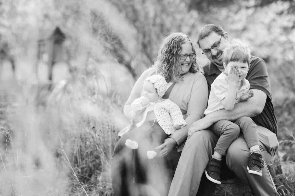 Black and white family portrait sitting outdoors at Quiet Waters Park. A woman holds a baby, and a man holds a young child who is playfully covering their face. Trees and grass are in the background, capturing a serene moment of joy.