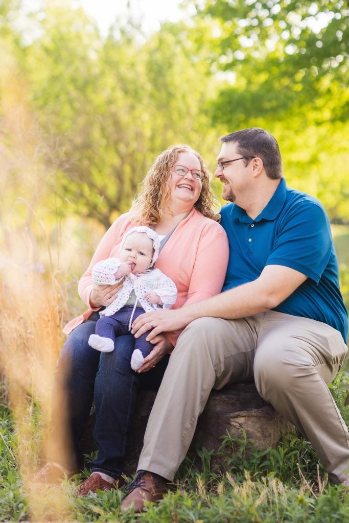 A couple sits on a rock at Quiet Waters Park, smiling at each other while holding a baby. The background features green foliage and sunlight, capturing the essence of a perfect family portrait.