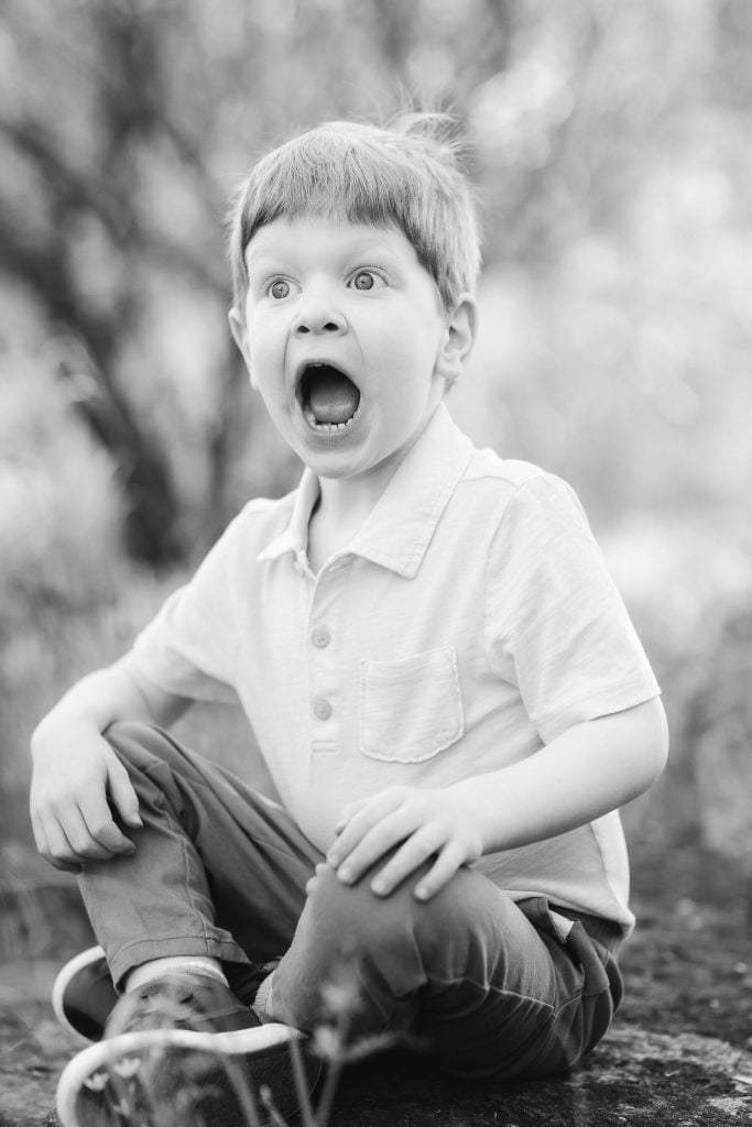 A family portrait captures a young boy with short hair, wearing a polo shirt and pants, sitting at Quiet Waters Park with his mouth wide open in an expression of surprise or excitement.