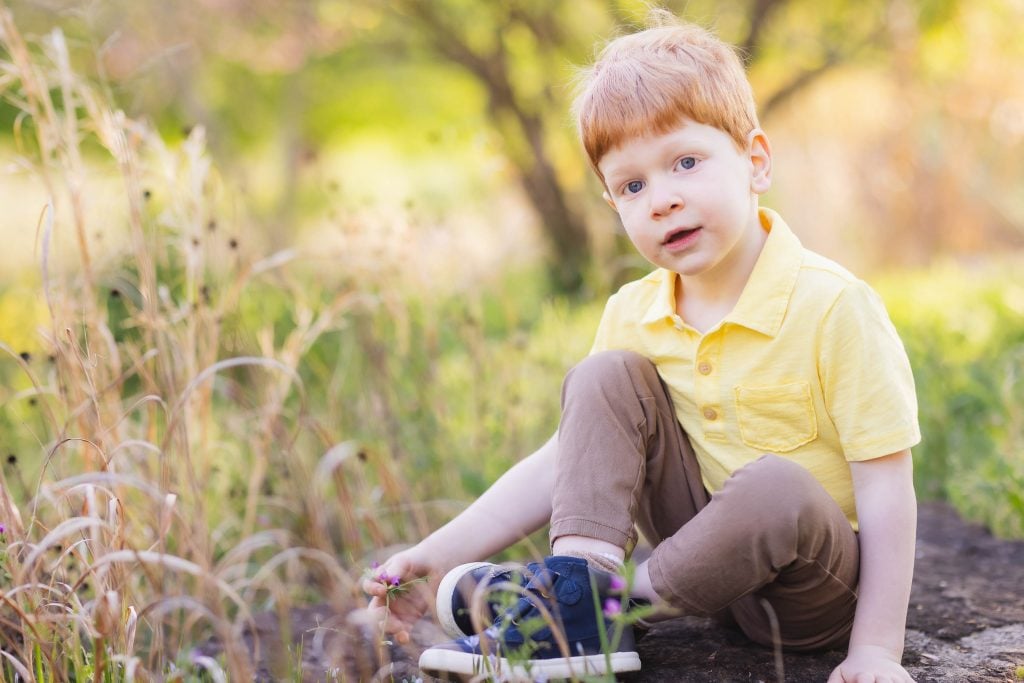 A young boy with red hair and a yellow shirt sits on a rock in the grassy, sunlit area of Quiet Waters Park, looking at the camera for a serene portrait.