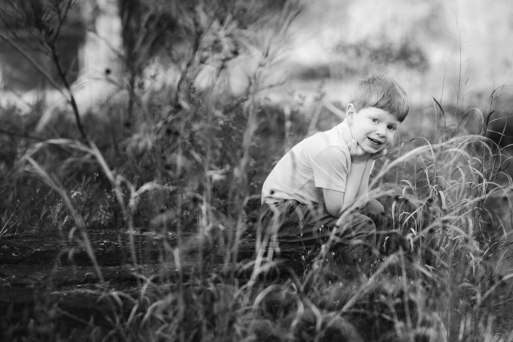 A young boy with short hair sits amidst tall grass in Quiet Waters Park, resting his head on his hands, and smiles at the camera. The scene is in black and white, capturing a serene family moment.