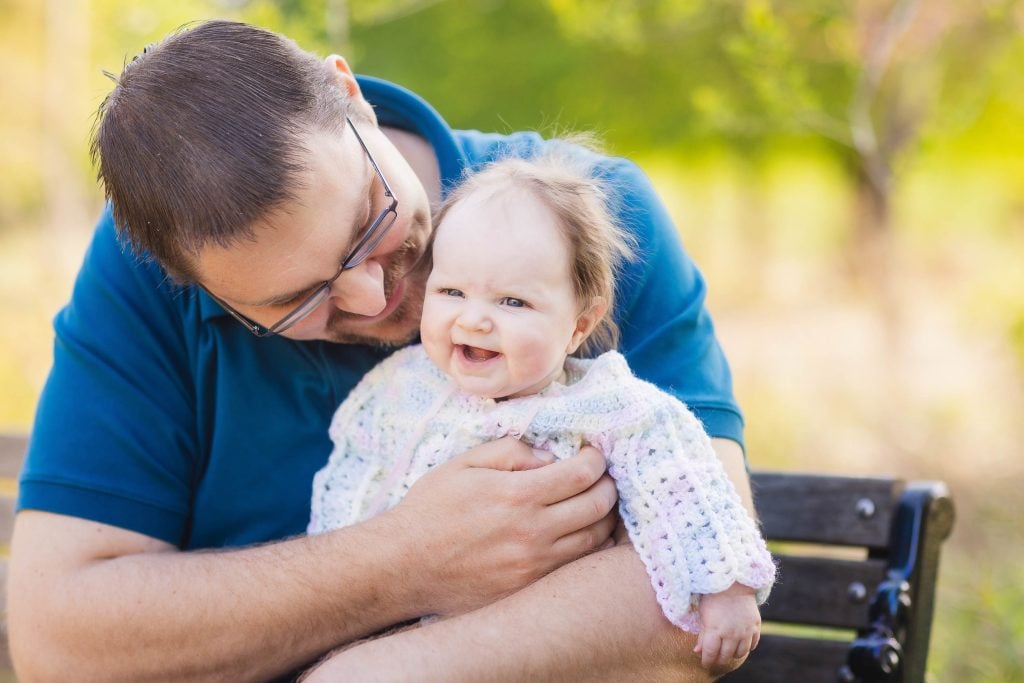 A man in a blue shirt holds a smiling baby wearing a knitted sweater while sitting on a bench at Quiet Waters Park, creating the perfect family portrait.