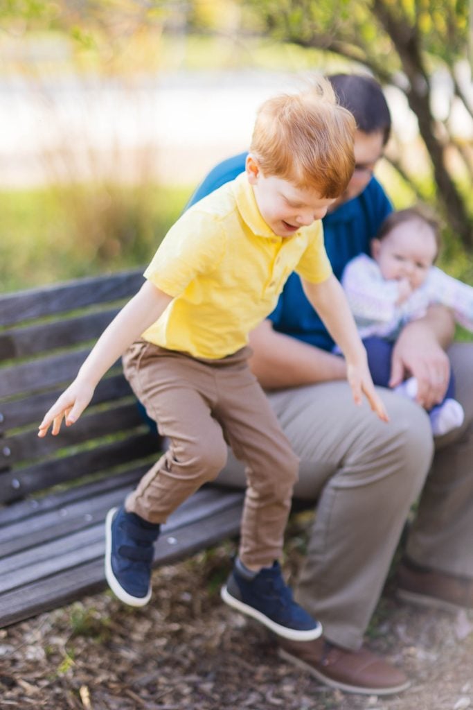 A young boy in a yellow shirt and brown pants jumps off a park bench at Quiet Waters Park while an adult holding a baby sits on the bench in the background, capturing the essence of family moments.