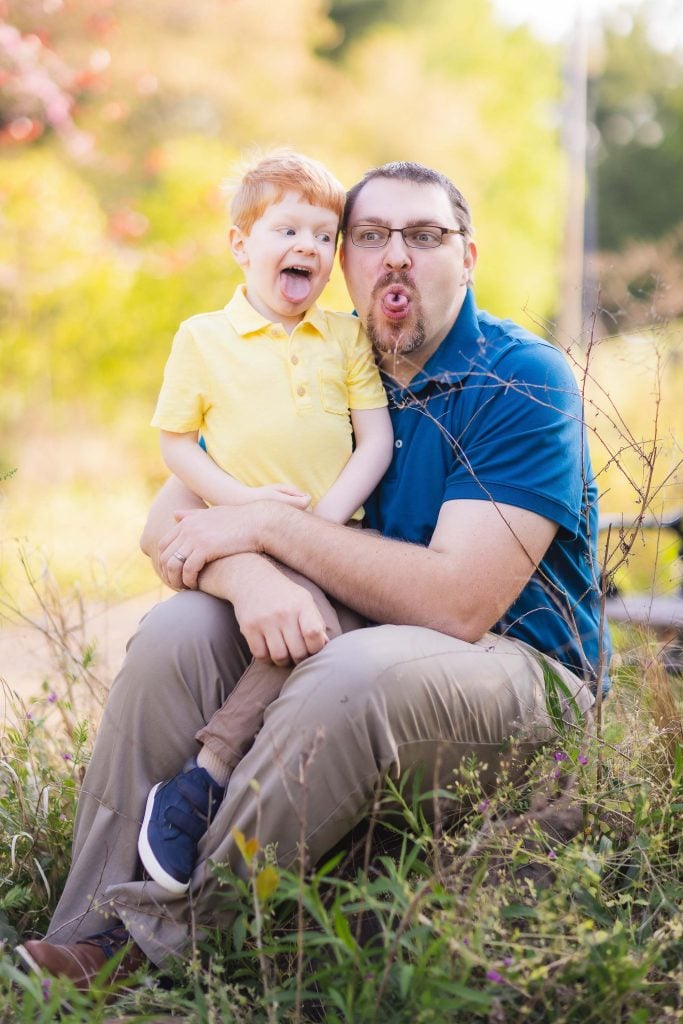 A man and a child sit together outdoors at Quiet Waters Park, making silly faces with tongues sticking out. The man wears glasses and a blue shirt; the child has red hair and wears a yellow shirt. Lush greenery surrounds their delightful family moment, creating a perfect portrait setting.