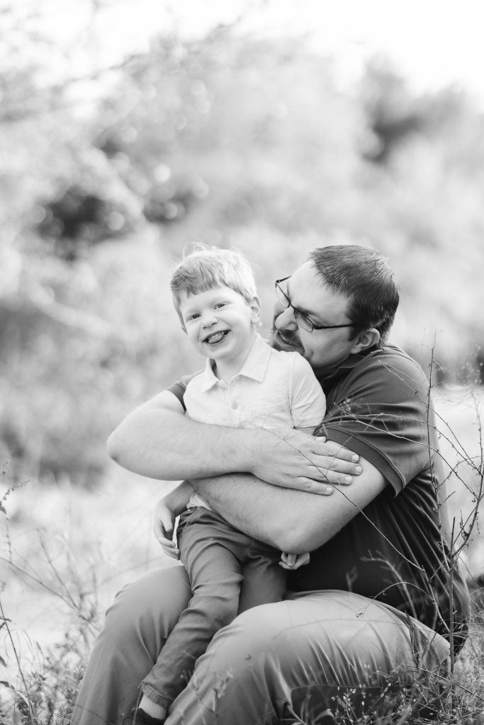 A man wearing glasses embraces and smiles at a young child with short hair, who is also smiling. They are sitting outdoors in a grassy area at Quiet Waters Park. The image, capturing a tender family moment, is in black and white.