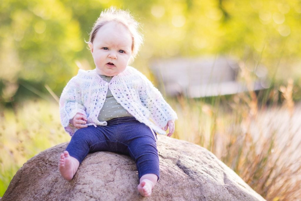 A baby wearing a crochet sweater and blue pants sits on a large rock in Quiet Waters Park, surrounded by greenery, looking at the camera with a slight frown. This charming portrait captures the serene family moment perfectly.