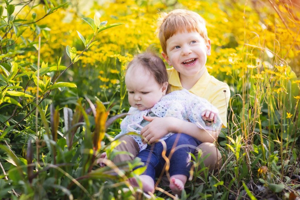 In Quiet Waters Park, a young child sits in a field of yellow flowers, holding a baby sibling on their lap. Both children are surrounded by lush green foliage, creating the perfect setting for timeless family portraits.