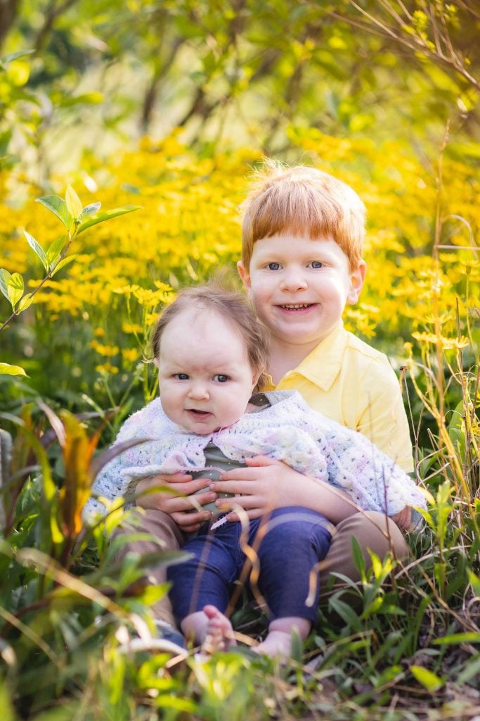 A young boy and a baby girl sit together in a grassy area surrounded by yellow flowers at Quiet Waters Park. The boy is holding the baby in his lap, both smiling at the camera. This serene family portrait captures a precious moment of sibling love and natural beauty.