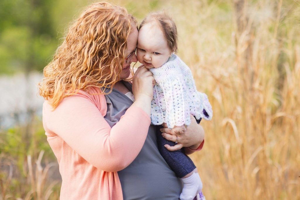 A woman with curly red hair holds a baby dressed in a white crocheted sweater, standing outdoors near tall grass at Quiet Waters Park.