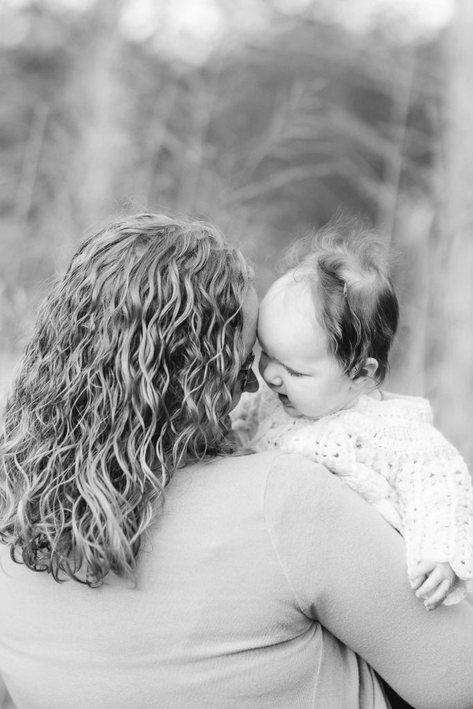 A person with curly hair holds a baby close, their foreheads touching. The baby, dressed in a knit sweater, gazes at the person. This tender moment captures the essence of family portraits often seen at Quiet Waters Park.