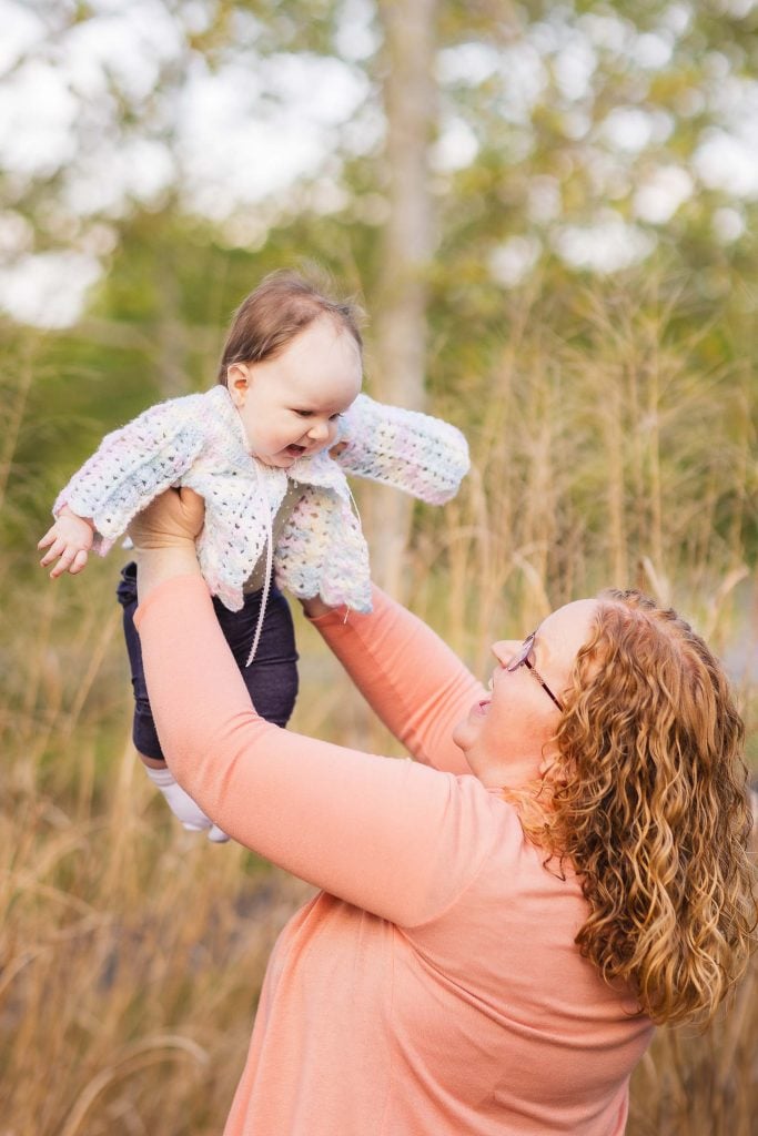 In Quiet Waters Park, a woman with curly hair lifts a baby wearing a knitted cardigan into the air, surrounded by tall grass, creating a beautiful family portrait.