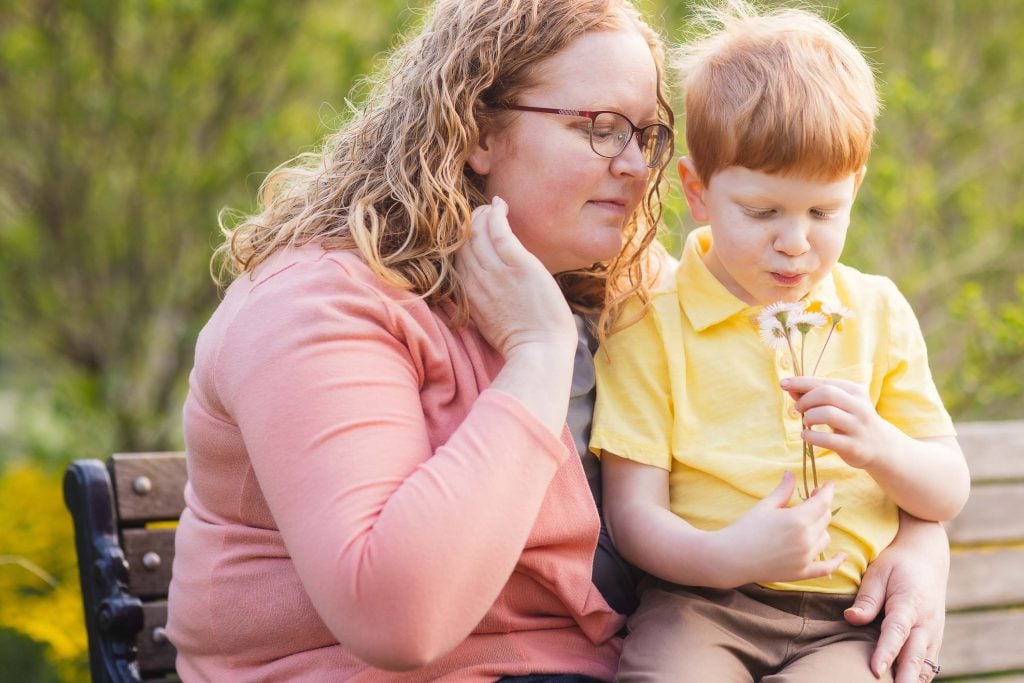 An adult with glasses and a child sit on a bench at Quiet Waters Park; the adult touches their face while the child looks at flowers they are holding, capturing a serene family moment.