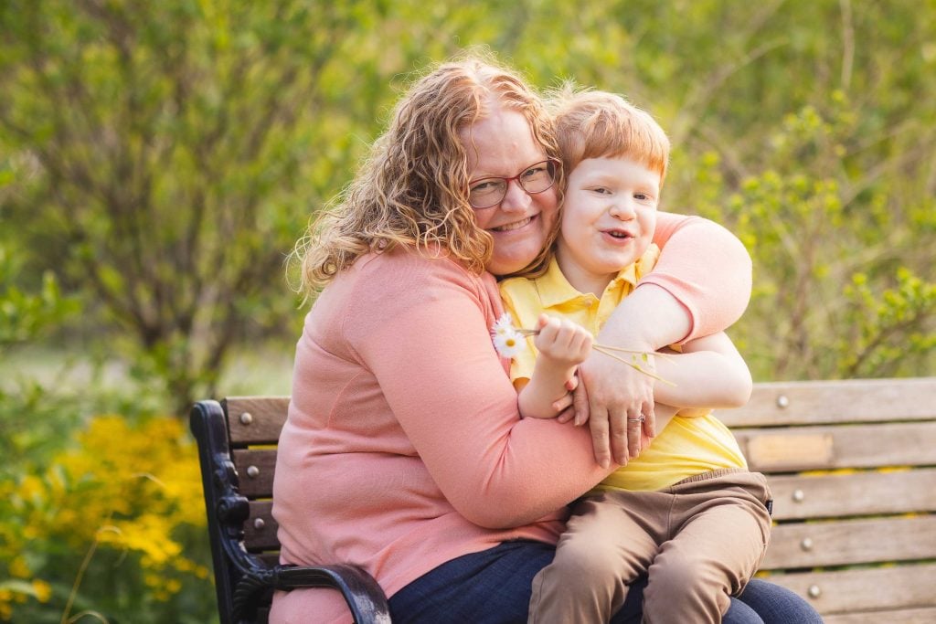 A woman wearing glasses hugs a smiling child holding a flower on a park bench with greenery in the background, capturing a heartwarming family moment in Quiet Waters Park.