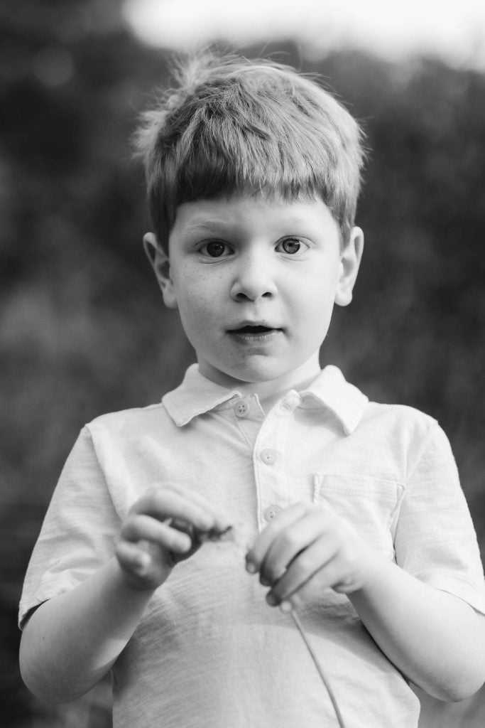 A black and white photo of a young boy with short hair wearing a lightcolored shirt. He looks at the camera while holding a small object, capturing a moment of innocence. The blurred background, possibly Quiet Waters Park, adds depth to this timeless family portrait.