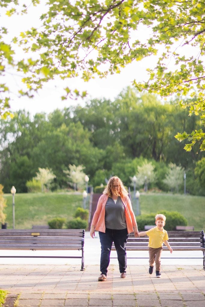 A woman and a young child walk handinhand on a paved path in Quiet Waters Park, with benches on either side and green trees in the background, capturing a serene family portrait.