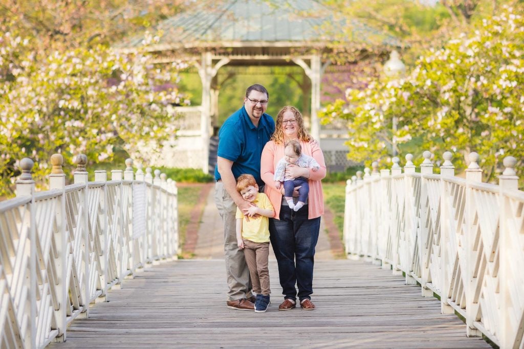 A family of four stands on a wooden bridge in Quiet Waters Park, with a gazebo and blooming trees in the background. The parents hold their two young children, smiling at the camera, creating a perfect family portrait.