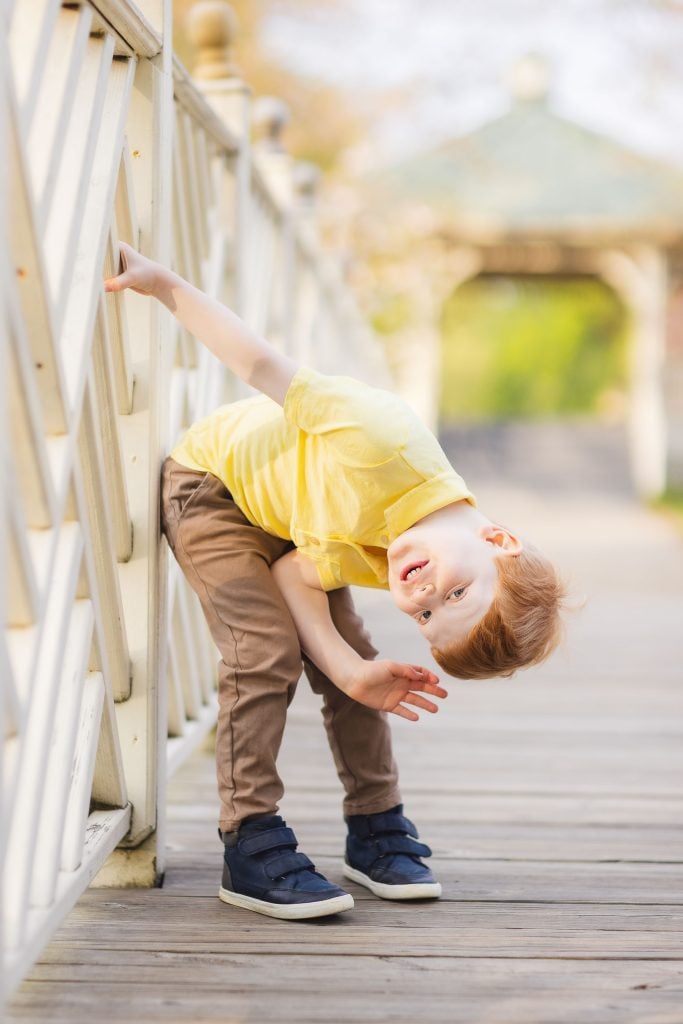 A young boy in a yellow shirt and brown pants leans sideways against a wooden fence, smiling and waving on a sunny day at Quiet Waters Park.