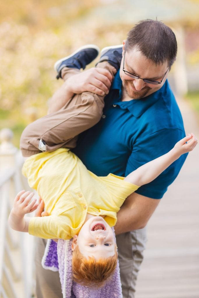 A man in a blue shirt is holding a laughing child upside down by the legs on a wooden walkway at Quiet Waters Park, with greenery in the background, capturing a joyful family moment.