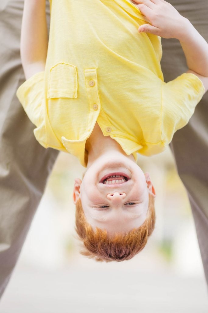 A child with red hair in a yellow shirt is hanging upside down, smiling broadly, capturing the joy of family moments at Quiet Waters Park. The background is slightly blurred, adding a dreamy quality to this playful portrait.