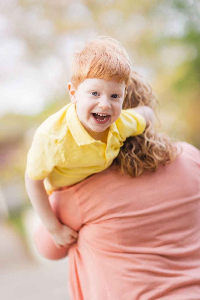 A child with red hair and a yellow shirt smiles while being carried on the back of a person wearing a pink top, outside at Quiet Waters Park with a blurred natural background, capturing the essence of family moments.