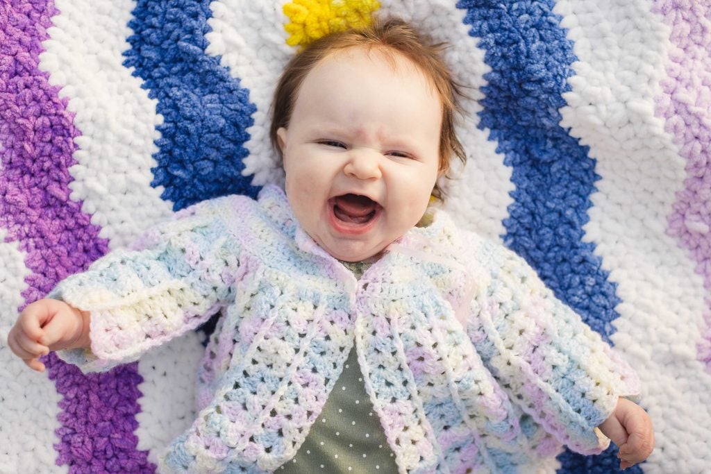 A baby with light hair smiles widely while lying on a crocheted blanket with blue, purple, and white stripes. The baby is wearing a crocheted sweater in pastel colors, creating a heartwarming family portrait reminiscent of moments cherished at Quiet Waters Park.