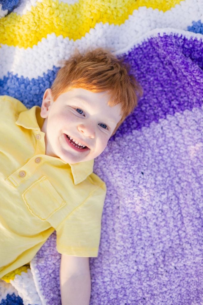 A smiling child with red hair lies on a colorful, striped blanket featuring yellow, white, blue, and purple sections, creating a pictureperfect moment for family portraits.