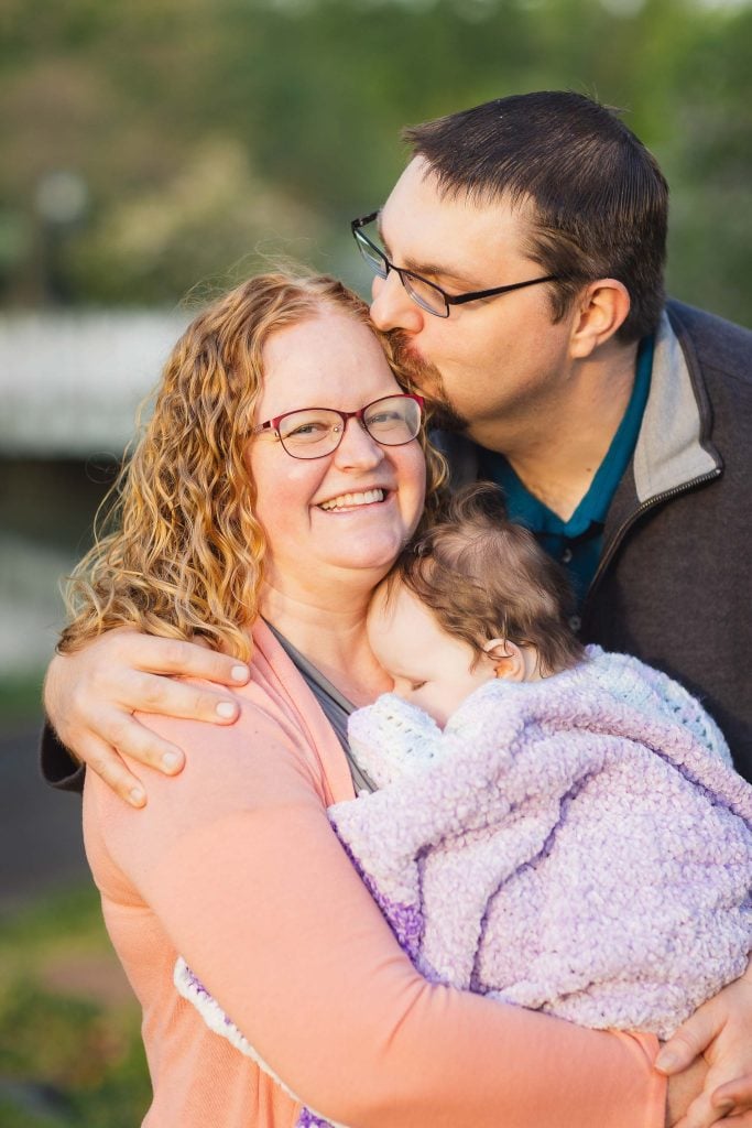 A family shares a tender moment as a man kisses a smiling woman on the forehead while she holds a baby wrapped in a purple blanket. This heartwarming scene is set outdoors at Quiet Waters Park, with lush green foliage providing the perfect backdrop for their portrait.