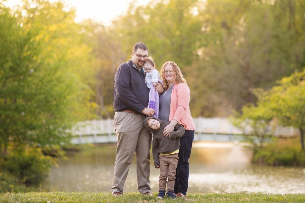 A family of four stands outdoors in front of a small lake and a bridge at Quiet Waters Park, with trees in the background. One child is held by an adult, while the other is playfully hanging upside down, making for perfect family portraits.