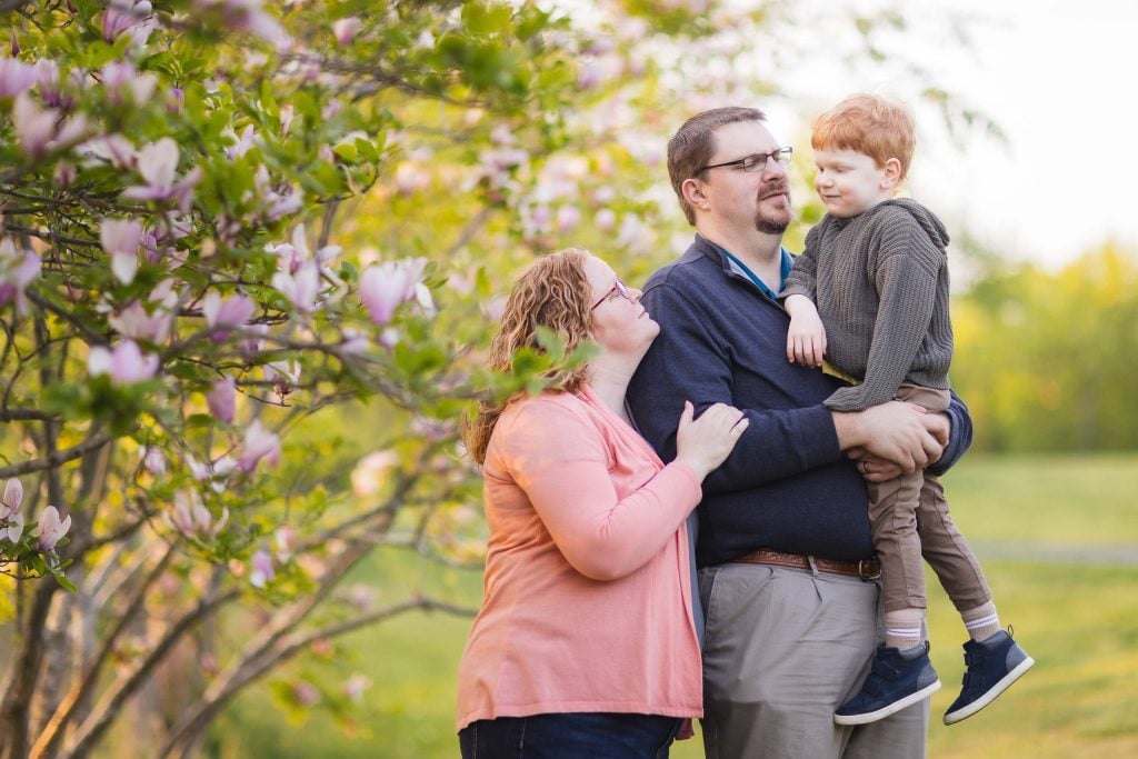 A man holds a young child while a woman stands beside them, looking up at the child. They are outdoors at Quiet Waters Park, surrounded by blooming trees—a perfect family portrait moment.