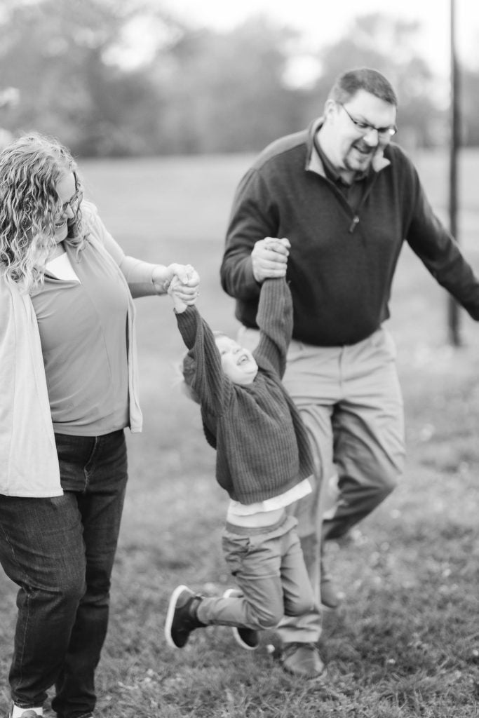 A child swings between two adults holding hands while walking in Quiet Waters Park. All are smiling, capturing a perfect family portrait. The image is in black and white.