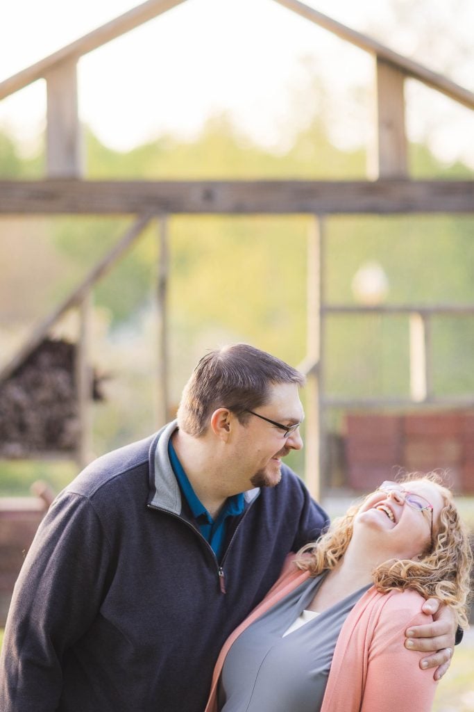 A man and a woman stand outdoors at Quiet Waters Park, sharing a joyful moment. The man has his arm around the woman as she laughs. A partially constructed wooden structure is in the background, giving the scene a timeless feel reminiscent of family portraits.