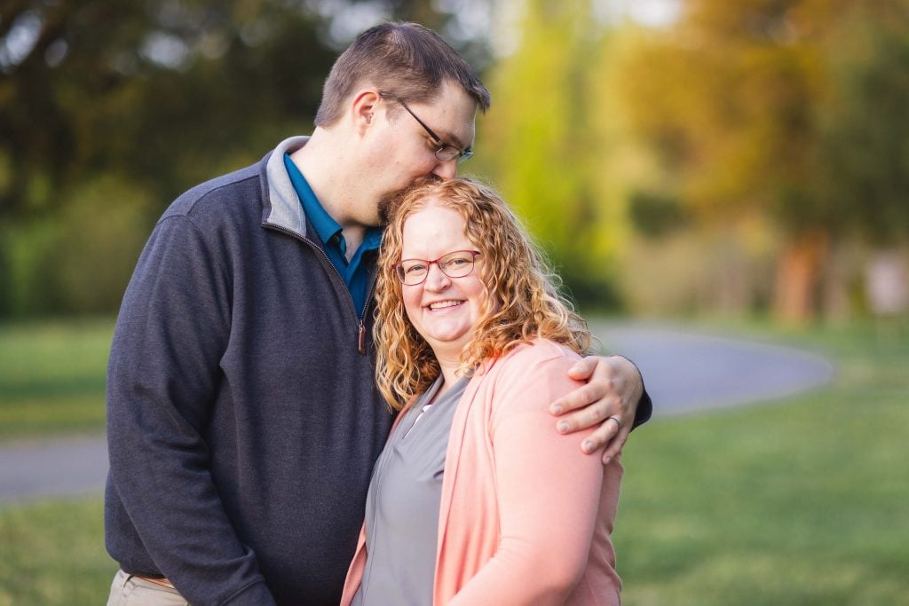 A man embraces and kisses a smiling woman on the head while standing outdoors at Quiet Waters Park, with a blurred background of trees and a pathway.