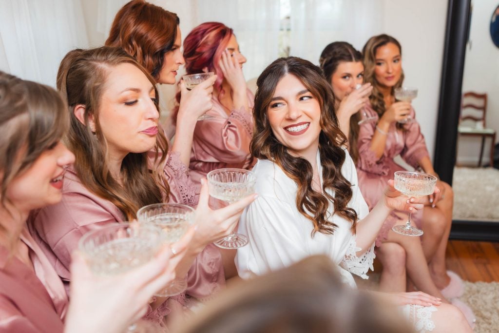 A bride and seven bridesmaids in matching robes smile and raise their glasses in a toast, sitting closely together in a bright room at The Barns at Hamilton Station, excitedly celebrating the wedding preparation.