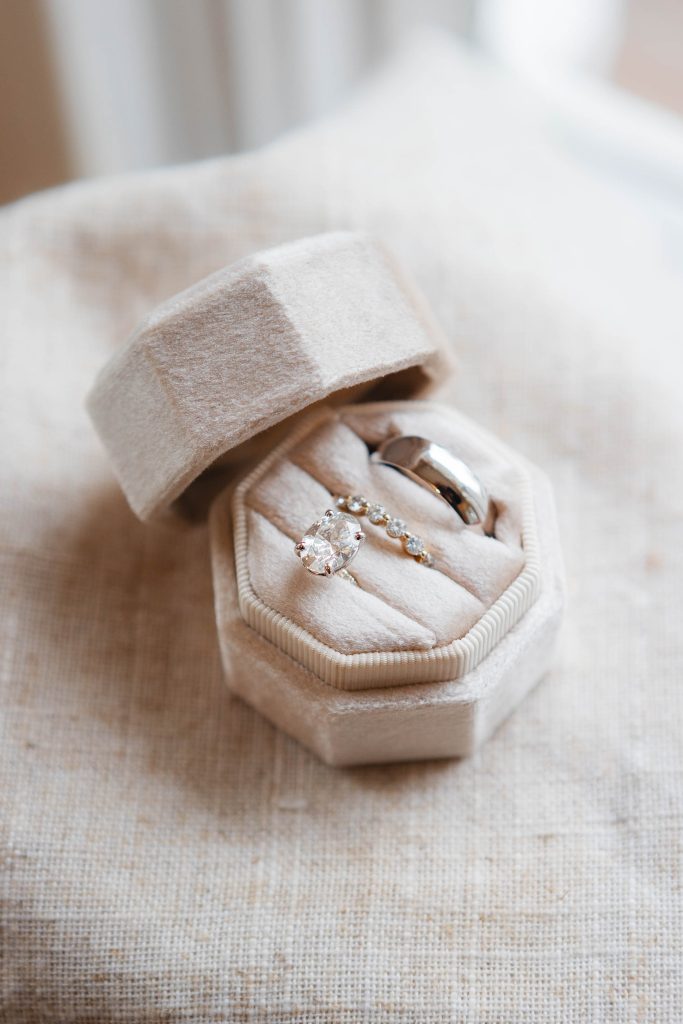 A beige velvet hexagonal ring box, perfect for a wedding at The Barns at Hamilton Station, holds two exquisite rings: one featuring a large ovalshaped gemstone and the other a plain band adorned with small, intricate details.