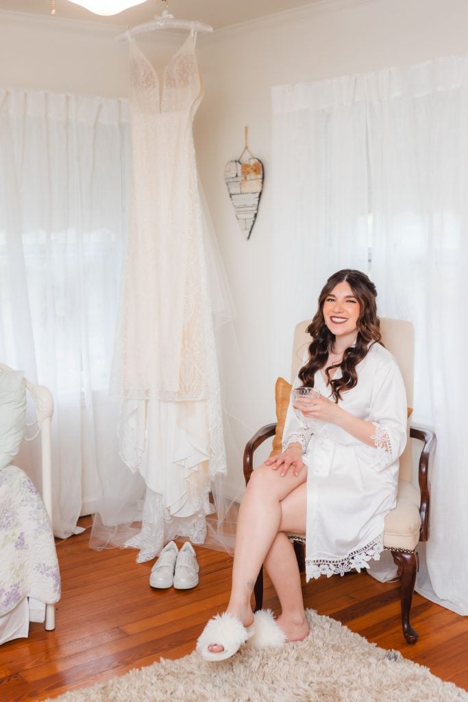 A woman in a white robe and fluffy slippers sits on a chair smiling, holding a drink. Behind her, in preparation for her big day at The Barns at Hamilton Station, a lace wedding dress hangs along with a pair of white sneakers.