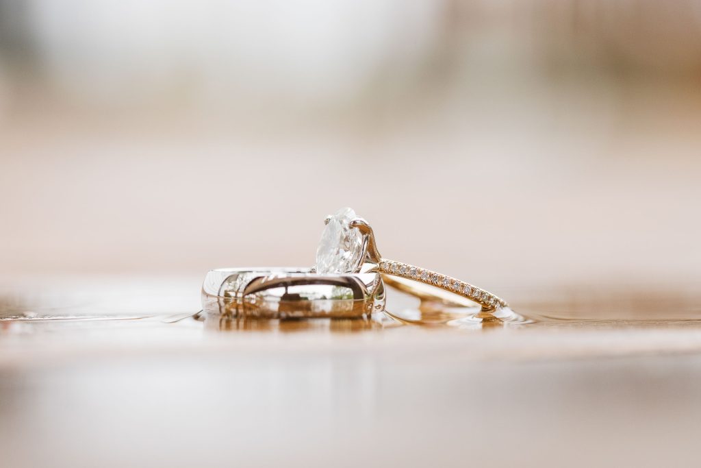 Closeup of two wedding rings, one with a large clear gemstone and a gold band with small inset stones, and the other a plain silver band, placed on a wet wooden surface at The Barns at Hamilton Station, showcasing exquisite details.