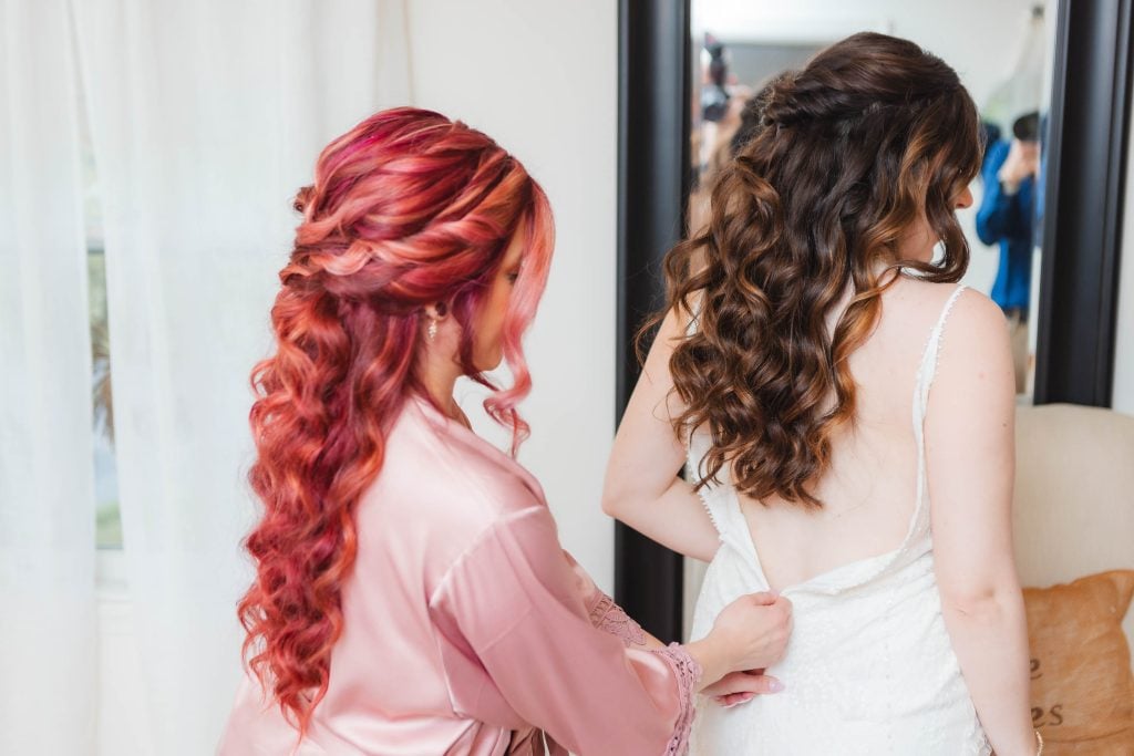 Two women with long curly hair, one with red and the other with brown, are seen from the back at The Barns at Hamilton Station. The woman with red hair is helping the woman in a white dress adjust her attire in front of a mirror, immersed in wedding preparation.