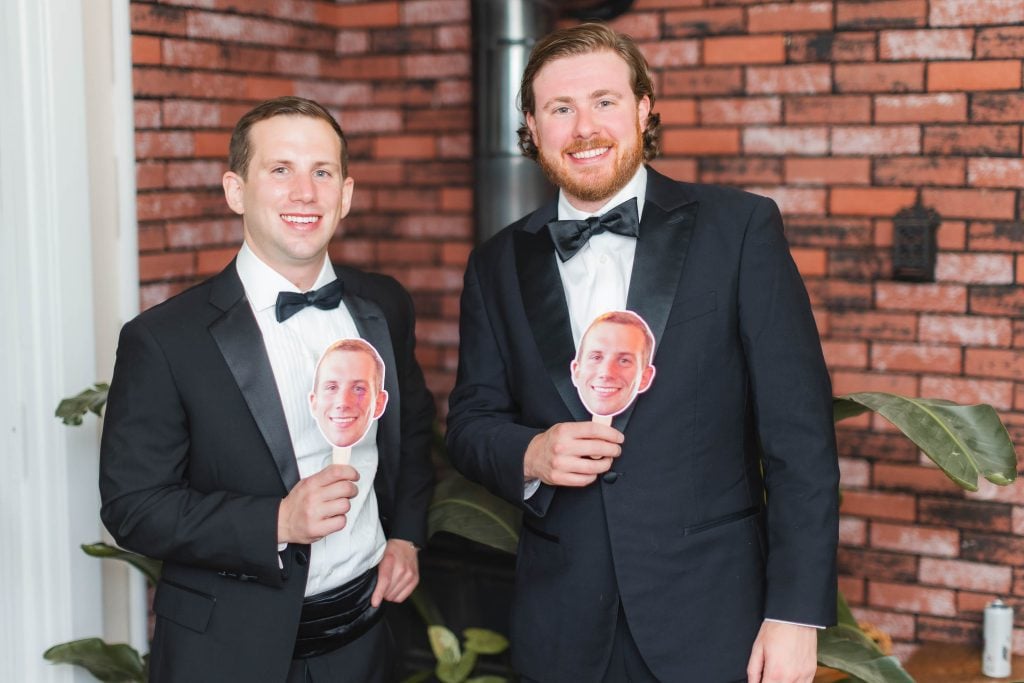 Two men in black tuxedos hold cutouts of the same smiling face in front of a brick wall, capturing the lighthearted moments of wedding preparation at The Barns at Hamilton Station.