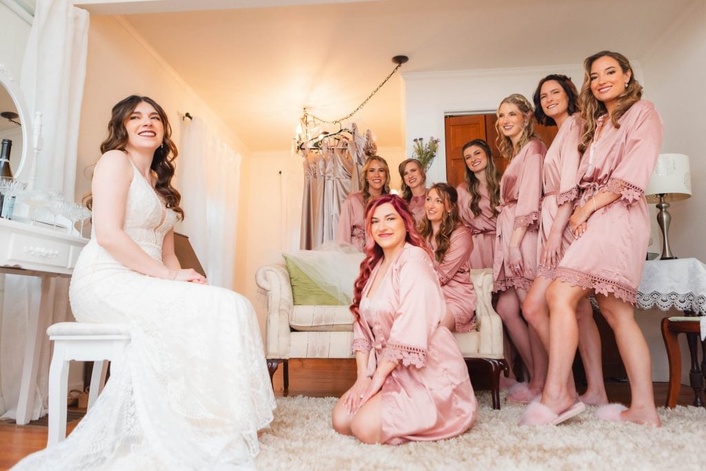 A bride in a white dress sits on a chair, the bridesmaids in pink robes gather around her, all smiling during the wedding preparation at The Barns at Hamilton Station. Soft lighting and a white rug complete the tender scene.