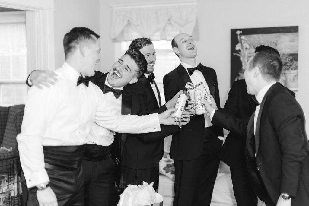 A group of five men in tuxedos share a moment of celebration and laughter, clinking aluminum cans together while preparing for the wedding at The Barns at Hamilton Station.