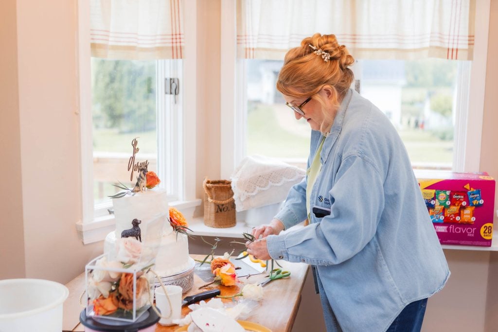 A person trims flowers and decorates a twotiered white wedding cake on a table near a window. The scene, set at The Barns at Hamilton Station, includes cake decorating tools, floral decorations, and a box of flavor packets in the background.