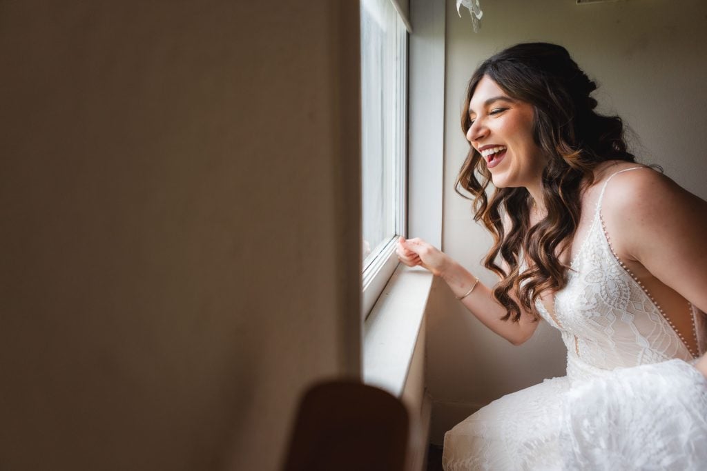 A woman in a wedding dress smiles and looks out of a window, with natural light illuminating the scene, as she enjoys a quiet moment amidst the wedding preparations at The Barns at Hamilton Station.