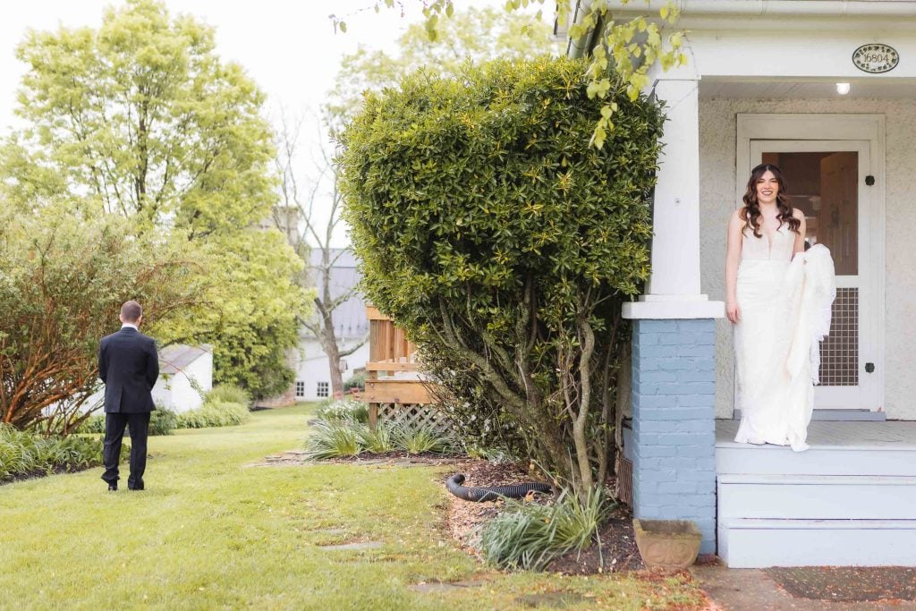 A bride in a white gown stands on the porch of The Barns at Hamilton Station, while a groom in a black suit, preparing for the wedding, faces away from her on the lawn, surrounded by greenery.